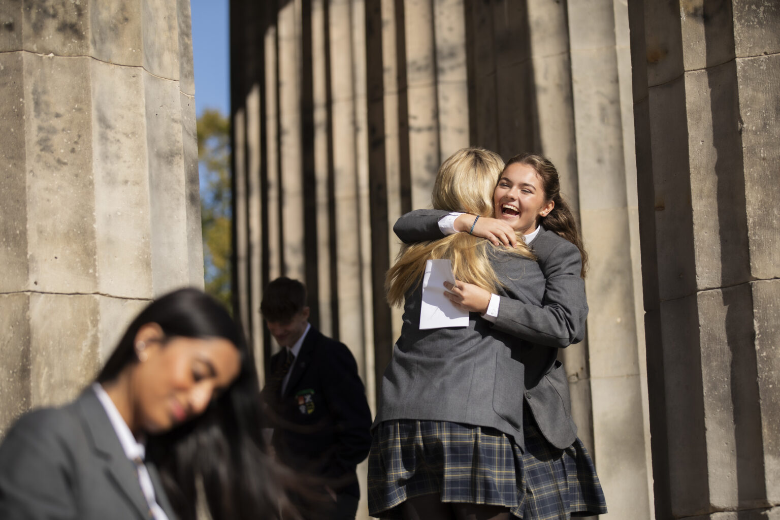 Believing. Pupils hugging each over and celebrating with documents in hand, at front of school by pillars.