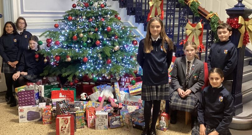 High School of Dundee pupils standing in hall by stairs, around lit up Christmas tree with presents for charity underneath the tree. Christmas decorations going up the banister of stairs.