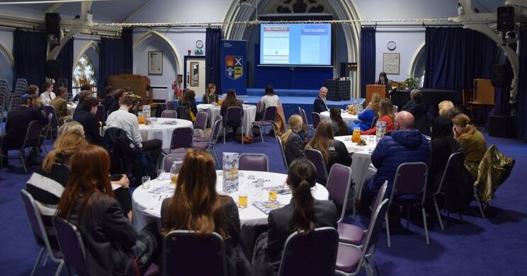 The Columns Happy 100th Edition! The celebratory 100th edition,HSD’s newsletter: by pupils; for pupils. Pupils and staff sat in a function room around tables giving presentations and celebrating.